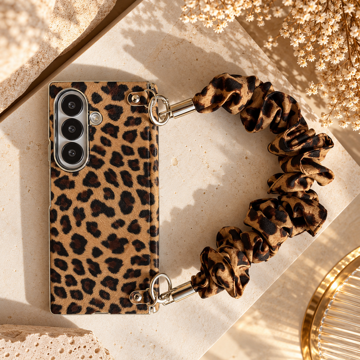 A phone case with a leopard print pattern and an attached scrunchie-style handle, styled on a beige stone surface with dried flowers and warm sunlight.